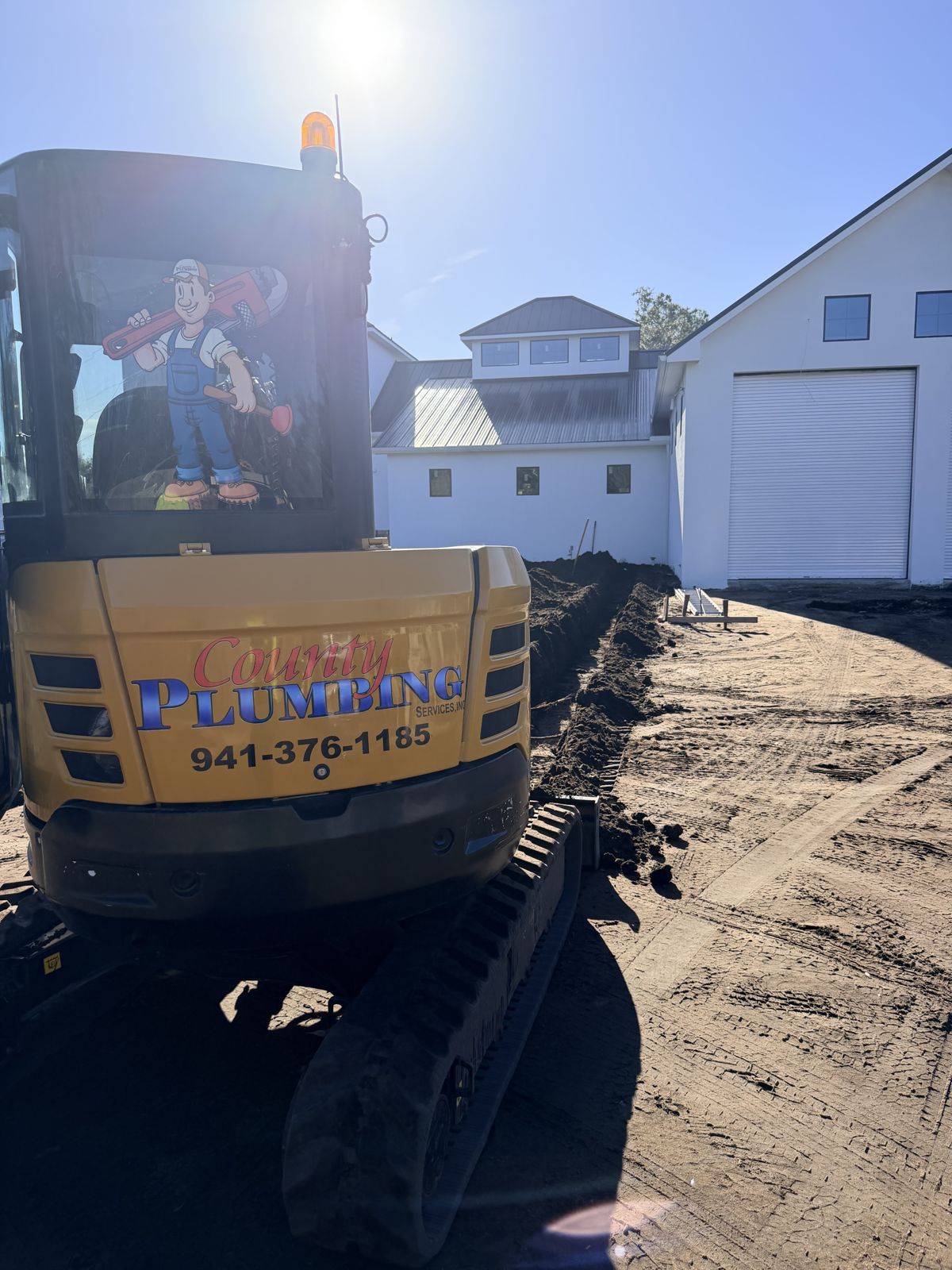 County Plumbing excavator with the plumber mascot and 941-376-1185 beside a fresh trench
